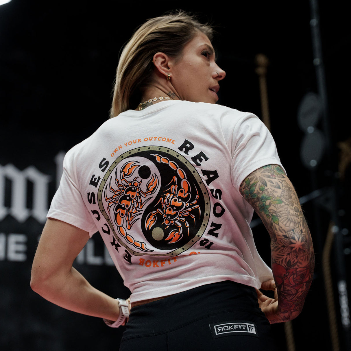 Female model in a gym wearing white crop top with 'Excuses or Reasons' graphic