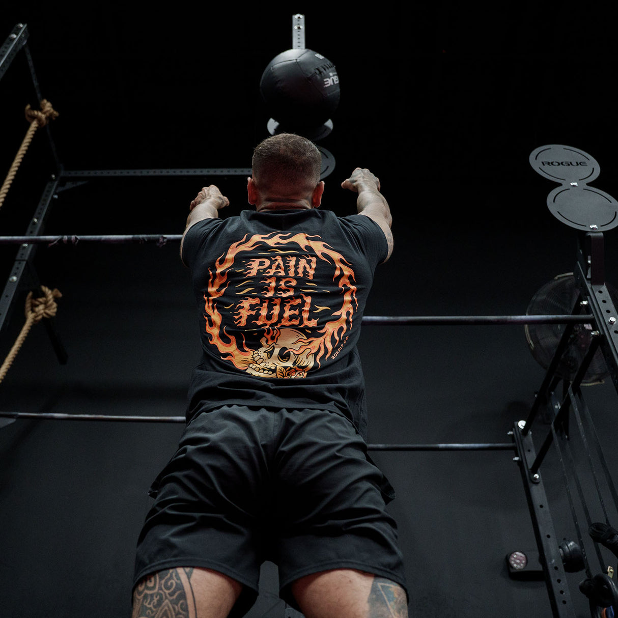 Male model doing a wall ball throw in a gym wearing Active Tee with 'Pain Is Fuel' graphic
