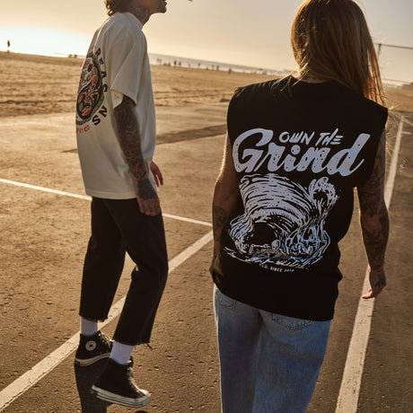 Female model outdoors wearing a sleeveless street tee with 'Own The Grind' graphic