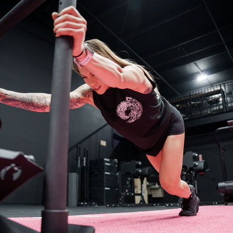 Female model pushing a weighted sled in a gym wearing sleeveless Street Tee with 'Own The Grind' graphic